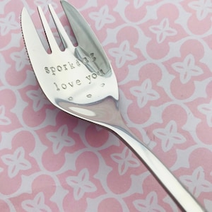 May include: A silver spork with the words "sporkling love you" engraved on the handle. The spork is on a pink and white patterned surface.