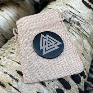 May include: A black round charm with a silver-colored Celtic knot design in the shape of a triangle. The charm is in a small burlap pouch.