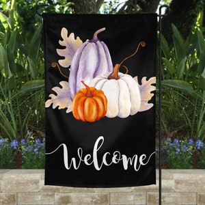 May include: A black welcome garden flag with a white welcome message and a watercolor illustration of three pumpkins. The pumpkins are purple, white, and orange.