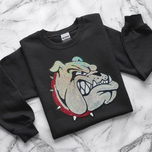 May include: Black sweatshirt featuring a sequined bulldog design. The bulldog is depicted with a fierce expression, surrounded by a red circle with white spikes. The design incorporates iridescent sequins for a shimmering effect. The sweatshirt has long sleeves.