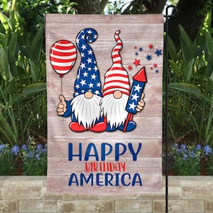 May include: A garden flag with two gnomes wearing red, white, and blue hats and outfits. The gnomes are holding a balloon and a firework. The flag has a wooden background with the text "Happy Birthday America".