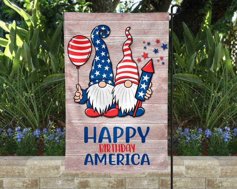 May include: A garden flag with two gnomes wearing red, white, and blue hats and outfits. The gnomes are holding a balloon and a firework. The flag has a wooden background with the text "Happy Birthday America".