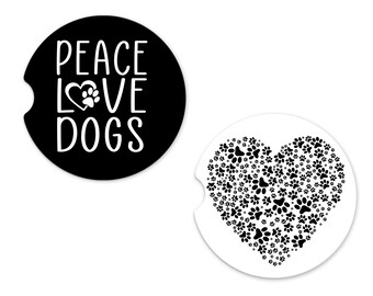 Puede incluir: Dos posavasos redondos en blanco y negro. Un posavasos tiene el texto "Peace Love Dogs" con un corazón y una huella de pata. El otro posavasos tiene forma de corazón hecho de huellas de pata.