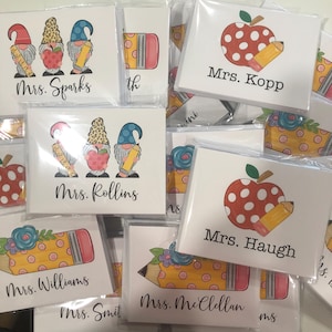 May include: A stack of white cards with different teacher names and illustrations. The illustrations include gnomes, pencils, apples, and flowers. The cards are packaged in clear plastic sleeves.