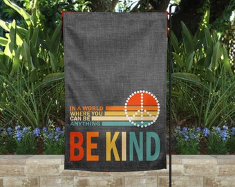 May include: A garden flag with a dark gray background and the words "BE KIND" in colorful letters. The flag also features a peace sign with a retro design and the phrase "IN A WORLD WHERE YOU CAN BE ANYTHING".
