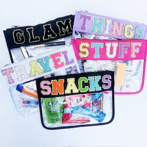 May include: Four clear plastic zippered pouches with different colored fabric tops. The tops have glitter letters spelling out "GLAM", "THINGS", "TRAVEL STUFF", and "SNACKS".