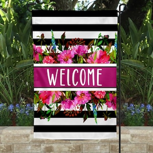 May include: A garden flag with a black and white striped background, featuring a vibrant floral design in pink, red, and blue. The word "WELCOME" is displayed in white on a magenta banner.