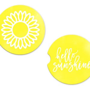 May include: Two yellow round coasters with white designs. One coaster features a sunflower design and the other coaster says "hello sunshine" in white cursive.