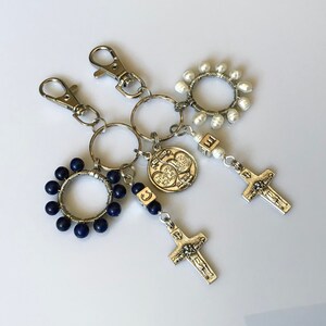 May include: A set of keychains with religious themes. The keychains feature a silver cross, a silver charm with two hearts, a blue rosary, and a white pearl rosary.