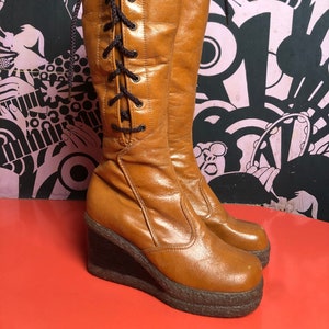 May include: Brown leather knee-high boots with a wedge heel and lace-up front. The boots have a shiny finish and a slightly pointed toe.
