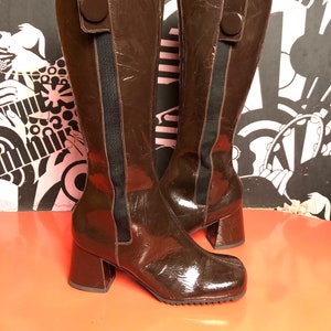 May include: A pair of brown leather knee-high boots with a chunky heel and a button detail on the side. The boots have a glossy finish and a black elastic panel on the back.