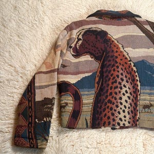 May include: A tapestry jacket featuring a detailed cheetah design. The jacket showcases a cheetah with brown spots, set against a backdrop of a savanna landscape in shades of blue, brown, and beige. The jacket has a collar and long sleeves.