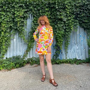 Pode incluir: Um vestido de manga curta com um padrão geométrico vibrante em tons de laranja, amarelo, rosa e branco. O vestido tem gola alta, uma carcela de botões na frente e uma bainha reta que cai acima do joelho, combinado com sandálias castanhas.
