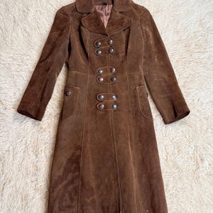 May include: A brown suede coat with a double-breasted design. The coat features large silver-colored buttons, a notched collar, and two front pockets. The coat is knee-length and has long sleeves.