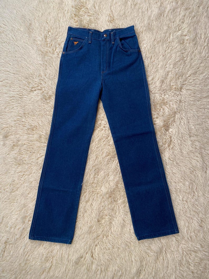 May include: A pair of blue denim jeans with a high waist and straight leg. The jeans have a classic five-pocket design, a button and zipper closure, and a small embroidered logo on the front pocket. The jeans are made of a sturdy denim fabric.