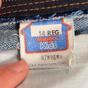May include: Close-up of a blue and white tag from a pair of Wrangler Kids jeans. The tag reads "14 REG" and features the Wrangler Kids logo. The tag also includes the code "47W06WD" and "Care Instructions-Over."