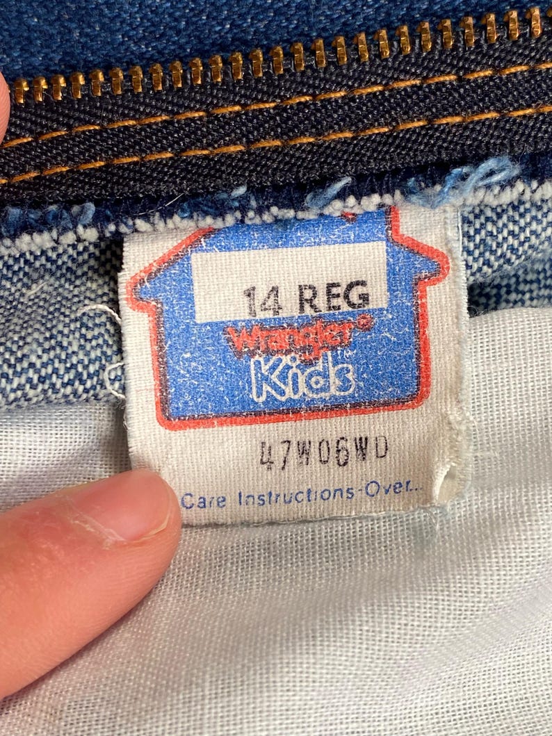 May include: Close-up of a blue and white tag from a pair of Wrangler Kids jeans. The tag reads "14 REG" and features the Wrangler Kids logo. The tag also includes the code "47W06WD" and "Care Instructions-Over."