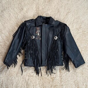 May include: A black leather jacket featuring long fringe detailing on the sleeves and front panels. The jacket includes a Bugs Bunny character patch on the upper chest, braided leather accents, and two circular silver-toned metal concho embellishments.