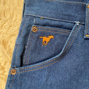 May include: Close-up of blue denim jeans with orange stitching. The jeans feature a small pocket with an embroidered orange horse logo. The jeans have metal button closures and a classic five-pocket design.