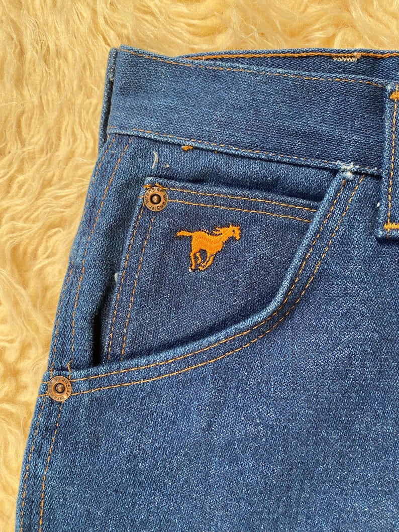 May include: Close-up of blue denim jeans with orange stitching. The jeans feature a small pocket with an embroidered orange horse logo. The jeans have metal button closures and a classic five-pocket design.