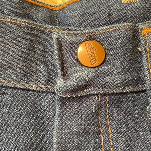 May include: Close-up of blue denim jeans with a copper-colored button. The button is embossed with the brand name "Wrangler". The jeans have orange stitching and a classic design.