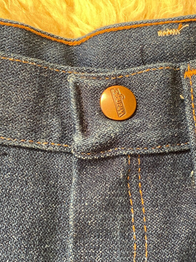 May include: Close-up of blue denim jeans with a copper-colored button. The button is embossed with the brand name "Wrangler". The jeans have orange stitching and a classic design.