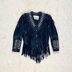 May include: Black leather jacket with fringe detailing, white bead accents, and a button-up front. The jacket features fringe along the chest and sleeves, with decorative beading around the collar and cuffs. The label reads "Sunriders".