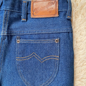 May include: Close-up of blue denim jeans with a brown leather patch that reads "Wrangler". The back pocket has a unique design with orange stitching. The jeans are made of a sturdy denim material.
