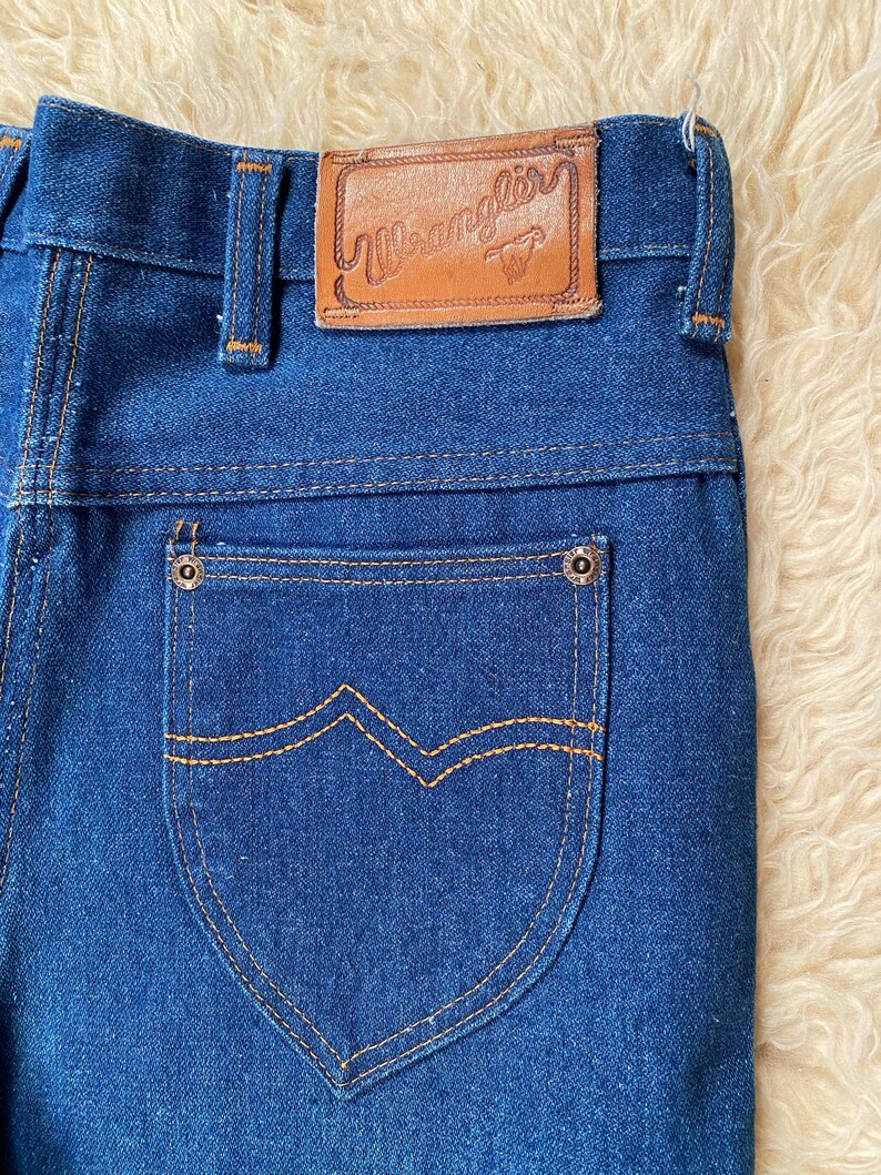 May include: Close-up of blue denim jeans with a brown leather patch that reads "Wrangler". The back pocket has a unique design with orange stitching. The jeans are made of a sturdy denim material.