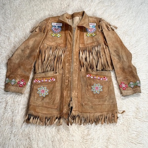 May include: A tan suede jacket with fringe detailing, pockets, and colourful floral embroidery. The jacket features a zip closure and decorative patches with text. The sleeves and hemline are adorned with long fringe.