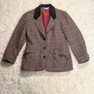 Peut inclure: Veste à carreaux marron et noir avec un col en velours noir et une doublure rouge. La veste a deux poches avant et trois boutons noirs. Les manches ont trois boutons aux poignets. La veste est posée sur une surface blanche et duveteuse.