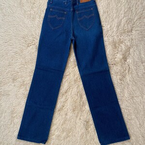 May include: Dark blue denim jeans with a classic five-pocket design. Features include a brown leather patch on the back and orange stitching. The jeans have a straight leg cut.