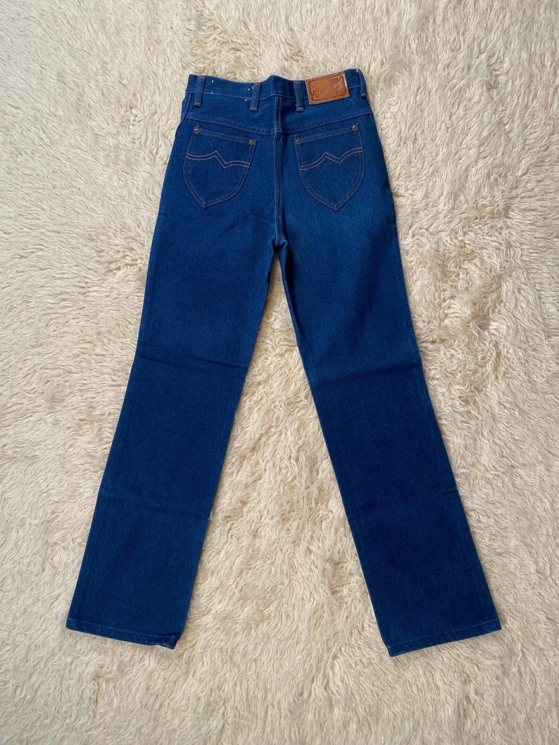 May include: Dark blue denim jeans with a classic five-pocket design. Features include a brown leather patch on the back and orange stitching. The jeans have a straight leg cut.