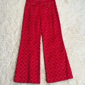 Puede incluir: Pantalones rojos brillantes de pierna ancha con un patrón blanco repetido y dos bolsillos delanteros. La tela tiene detalles de costura blancos. Los pantalones se muestran sobre una superficie blanca y esponjosa.