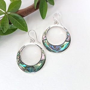 May include: A pair of silver hoop earrings with iridescent abalone shell inlays. The earrings have a circular design with a hook closure. The abalone shell displays a range of colors, including green, blue, and purple. The earrings are displayed on a white surface.