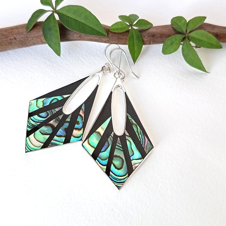 May include: A pair of diamond-shaped dangle earrings featuring iridescent abalone shell inlays set within black frames. The earrings have silver-toned metal borders and silver ear wires, with a central oval cutout at the top of each piece.