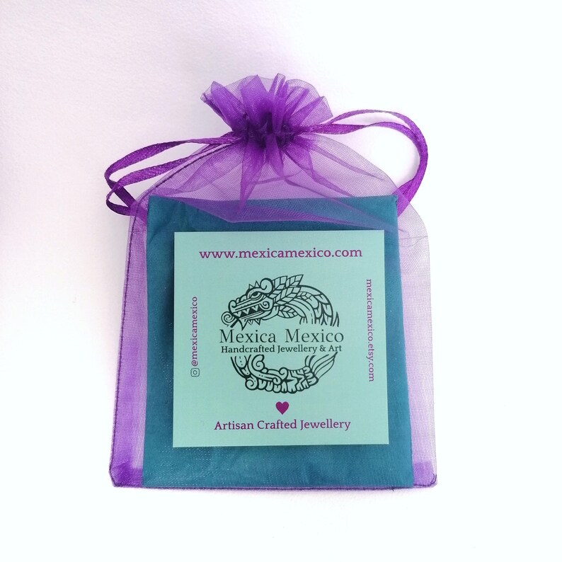May include: A purple organza gift bag with a drawstring closure, containing a teal-colored card and a teal fabric. The card features the text "Mexica Mexico Handcrafted Jewellery & Art" and a stylized dragon design.