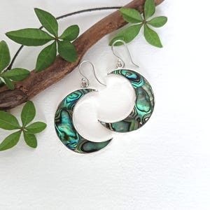 Può includere: Un paio di orecchini pendenti a forma di mezzaluna con intarsi in conchiglia di abalone iridescente incastonati in una cornice di metallo argentato. Ogni orecchino pende da un semplice gancio argentato, mostrando motivi vorticosi verdi e blu.