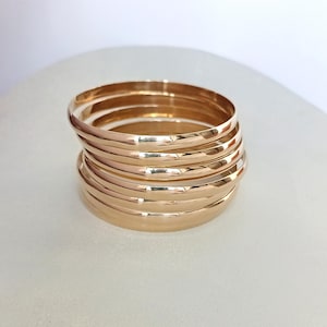Könnte beinhalten: Ein Stapel aus sieben polierten Armreifen in Roségold-Optik. Jeder Armreif hat eine glatte, abgerundete und reflektierende Oberfläche. Die Armreifen sind in einer vertikalen Säule angeordnet und zeigen ihre gleichmäßige Kreisform.