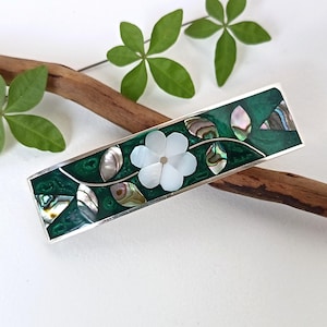 May include: A rectangular barrette with a silver frame. It features a dark green background with a floral design. The barrette has a white flower in the centre and iridescent abalone shell accents. The leaves and flower are made of mother of pearl.