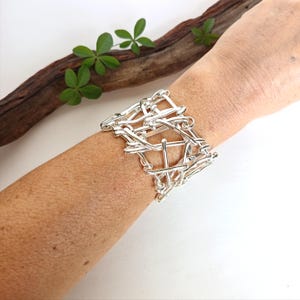May include: A wide cuff bracelet constructed from multiple interconnected silver-toned metal wires, forming an open, lattice-like structure that wraps around the wrist. The design features a series of overlapping and knotted metallic segments.