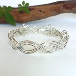 May include: A silver-coloured wire bracelet featuring a multi-strand design with a continuous wave pattern. The bracelet is constructed from several thin, polished metal wires that are woven together and secured at the ends with decorative wire wraps.