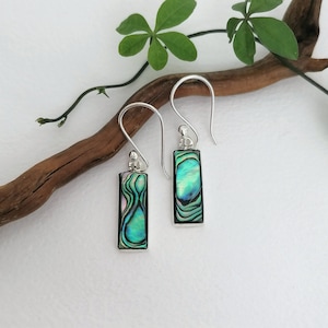 May include: Silver rectangular earrings with iridescent abalone shell inserts. The earrings feature a hook design and display green, blue, and purple colours. The earrings are presented on a white surface, with a wooden branch and green leaves.