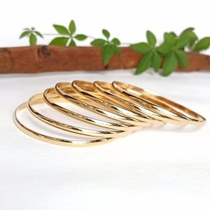 Pode incluir: Uma coleção de seis pulseiras douradas, cada uma com uma superfície lisa e polida. As pulseiras estão dispostas em semicírculo, mostrando seu design simples e elegante. Folhas verdes e um galho marrom ao fundo.