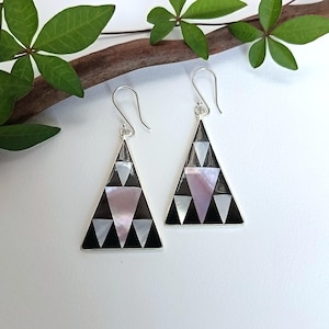 Könnte beinhalten: Ein Paar silberfarbene Dreiecks-Ohrringe. Jeder Ohrring hat ein geometrisches Design mit schwarzen, weißen und rosa Perlmutt-Inlays. Die Ohrringe hängen an einem einfachen Haken.