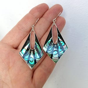 May include: A pair of diamond-shaped dangle earrings featuring a silver-toned frame with black enamel accents and iridescent abalone shell inlays. The earrings have a geometric design with a central cutout and silver ear wires for attachment.
