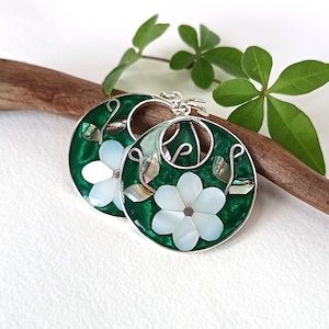 Puede incluir: Un par de pendientes circulares con un fondo de esmalte verde esmeralda intenso y una flor central blanca de seis pétalos. El diseño incluye detalles de concha iridiscente en forma de hojas y filigrana de metal en tono plateado.