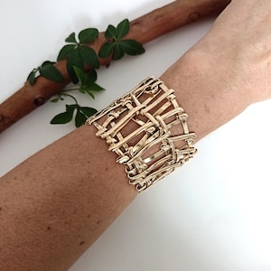 May include: A wide cuff bracelet crafted from polished gold-tone metal, featuring an intricate, open-work woven design with interlocking horizontal and vertical wire-like strands that create a structured, cage-like appearance around the wrist.