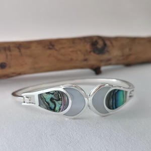 May include: A silver bangle bracelet with two oval-shaped inlays. The inlays are made of iridescent abalone shell and white mother of pearl.