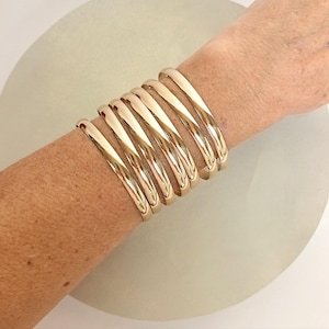 May include: A set of six polished gold-tone metal cuff bracelets worn together on the wrist. Each bracelet features a smooth, rounded, and reflective surface, creating a stacked appearance with uniform spacing between the individual metal bands.